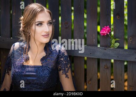 Belle fille en robe d'outremer glamour debout devant une clôture en bois avec une rose rouge attachée sur elle. Prêt pour sa soirée de bal. Banque D'Images