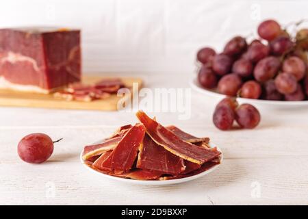 Émincé de jamon sur une plaque contre une grande pièce de jamon sur une planche à découper et un bouquet de raisins roses sur une table en bois blanc. Jamon espagnol traditionnel Serrano Banque D'Images