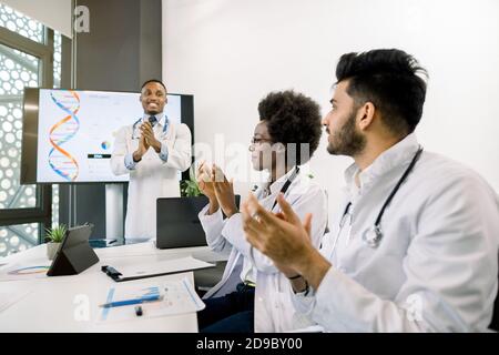 Un jeune médecin africain donne un discours réussi à l'équipe médicale, tout en se tenant devant un grand écran numérique avec des diagrammes et la structure de l'adn Banque D'Images