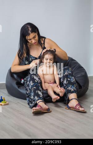 Une mère de Latina peigne sa petite fille de 18 mois assise sur un pouf, avec des jouets en bois à côté d'elle. Banque D'Images