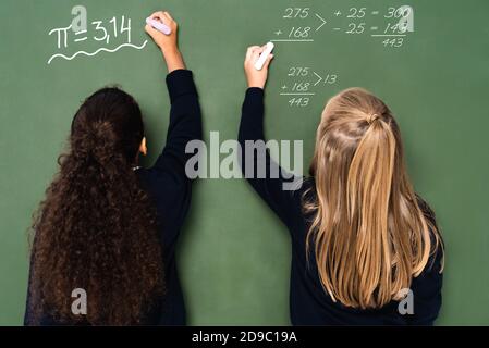 vue arrière de filles d'école multiculturelles écrivant des équations mathématiques sur tableau noir Banque D'Images