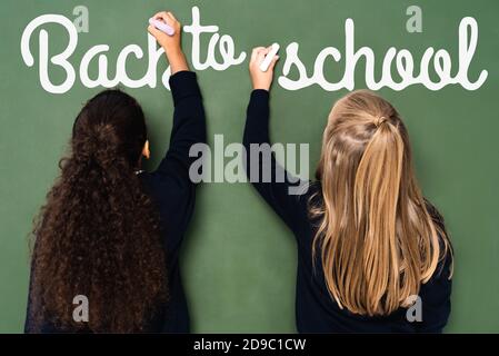vue arrière de filles d'école multiculturelles écrivant à l'école lettering sur tableau noir Banque D'Images