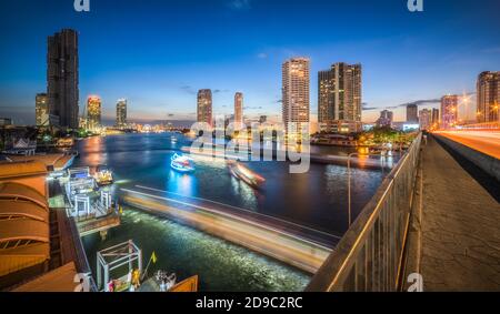 Gratte-ciels et pistes lumineuses de trafic intense sur la rivière Chao Phraya à Bangkok, Thaïlande, vu depuis le pont Taksin au crépuscule Banque D'Images