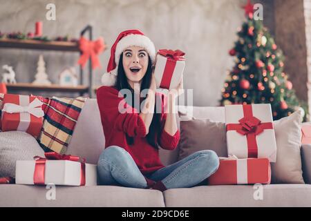 Photo de jolie fille assis surprise face ouverte bouche canapé porte-boîte cadeau vêtements chapeau de père noël rouge chandail jeans dans la décoration séjour à l'intérieur Banque D'Images