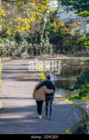 Un jeune couple bras dans le bras marchant le long d'une rivière sous les arbres d'automne lors d'une journée ensoleillée à Christchurch dans dorset. Banque D'Images