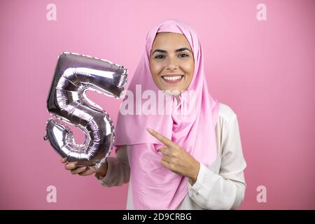 Jeune belle femme arabe portant le hijab islamique sur fond rose isolé souriant tenant un ballon numéro cinq. Concept de célébration Banque D'Images