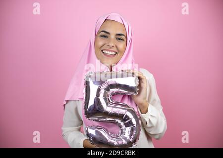 Jeune belle femme arabe portant le hijab islamique sur fond rose isolé souriant tenant un ballon numéro cinq. Concept de célébration Banque D'Images