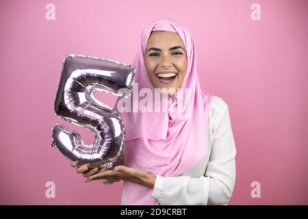 Jeune belle femme arabe portant le hijab islamique sur fond rose isolé souriant tenant un ballon numéro cinq. Concept de célébration Banque D'Images