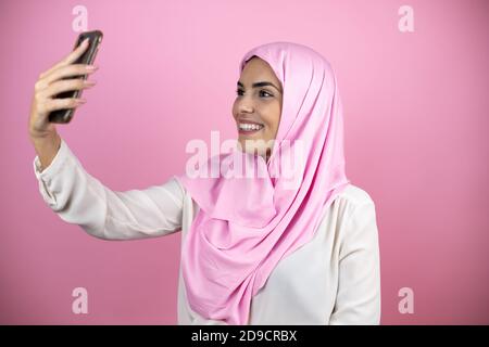 Jeune belle femme arabe portant le hijab islamique sur le rose isolé arrière-plan prenant un selfie avec son téléphone Banque D'Images