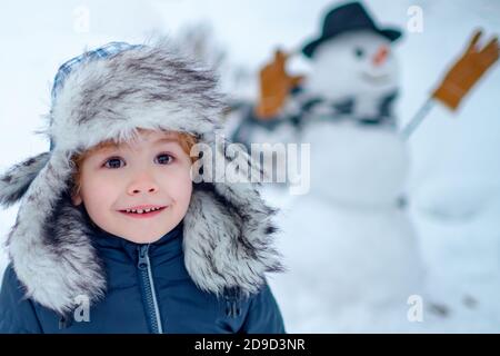 Bonhomme de neige et enfant drôle l'ami est debout dans un chapeau d'hiver et un foulard avec le nez rouge. Faire bonhomme de neige et l'hiver s'amuser pour les enfants. Drôle de garçon posant dessus Banque D'Images