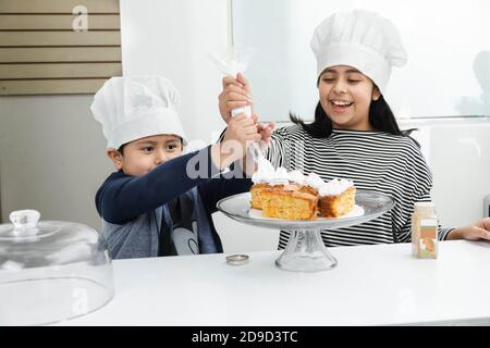 Enfants hispaniques décorant un gâteau avec de la crème fouettée - Latino frères et sœurs s'amuser tout en mettant de la crème fouettée sur le gâteau - les enfants cuisent Banque D'Images