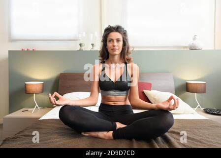 S'adapter une femme en bonne santé méditant dans la chambre assise sur le lit dans la posture de lotus face à l'appareil photo avec un un sourire serein Banque D'Images