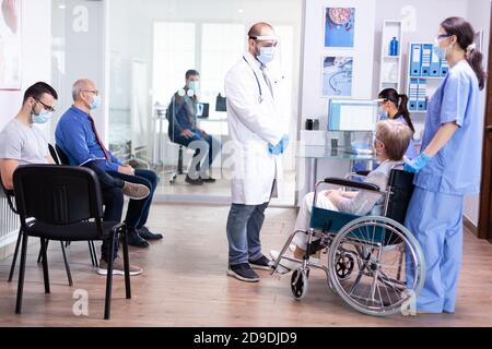Jeune médecin portant un masque facial contre le coronavirus parlant avec une femme handicapée en fauteuil roulant pendant l'examen contre le covid-19. Jeune homme en attente de consultation médicale. Système de soins de santé. Banque D'Images