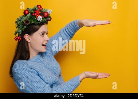 Une jeune femme choquée dans une couronne de Noël montrant quelque chose de grande taille regardant les mains, fille stupéfaite avec la bouche ouverte surprise avec une remise énorme Banque D'Images