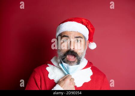 Portrait de l'homme dans le chapeau de Santa prend le masque médical facial de son visage. Célébration de Noël pendant la pandémie du coronavirus Banque D'Images