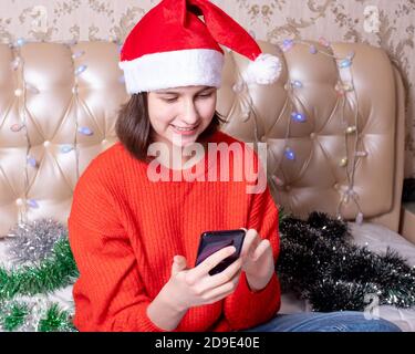 Une fille portant un chapeau de père noël sourit et clique sur son smartphone pour faire des achats en ligne ou discuter avec des amis en ligne Banque D'Images