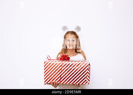 Une belle petite fille tenant une boîte cadeau de Noël à rayures rouges avec un ruban et un noeud dans ses mains. Il y a des flocons de neige sur la tête. Isolé sur Banque D'Images