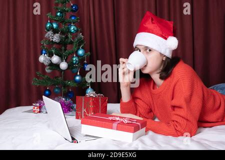 Une jeune femme dans un chapeau de père Noël et un chandail rouge boit du café, se reposant avec son ordinateur portable, allongé sur le lit Banque D'Images