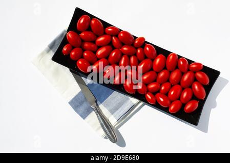 Tomates cerises rouges sur une assiette rectangulaire noire de style oriental et un couteau en acier sur une serviette. Arrière-plan blanc. Copier l'espace pour le texte. Banque D'Images