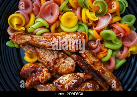 Viande de porc frite dans une sauce caramel servie avec du multi pâtes colorées sur plaque céramique noire, cuites à plat Style directement d'en haut. Délicieux barbecue Banque D'Images