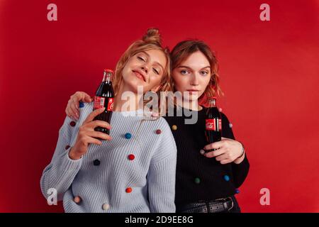 Deux jolies petites amies tiennent des bouteilles de coke. Une fille a mis sa main sur l'épaule d'une autre. Isolé sur un arrière-plan rouge. Banque D'Images