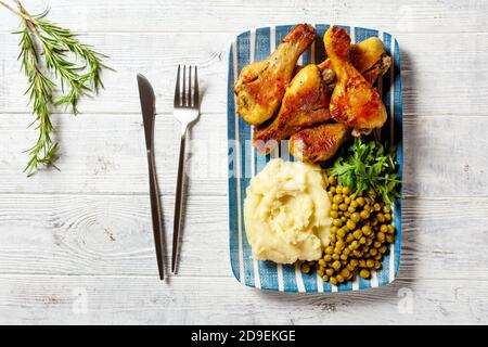 Dîner facile : pilons de poulet cuits servis sur une assiette avec des arugula fraîches, une purée de pommes de terre et des petits pois sur un fond de bois blanc avec des couverts, Top vie Banque D'Images