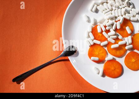 Capsules blanches et tranches de persimmon orange sur une plaque ovale blanche et une petite cuillère en métal sur fond orange. Suppléments et minéraux Banque D'Images