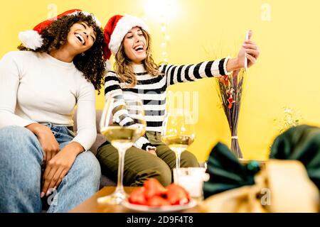 Vacances en famille avec diversité: Belle blonde prend un selfie à la maison avec sa petite amie hispanique noire portant chapeau de Santa - les gens modernes différents Banque D'Images
