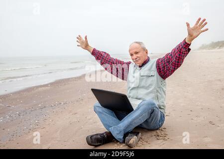 Un vieil homme heureux assis sur la plage avec un ordinateur portable et levant les mains.travailler pendant la quarantaine, pandémie Banque D'Images