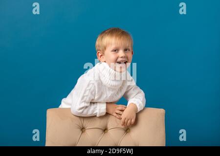 Un garçon joyeux, un enfant blond dans un pull blanc chaud, se tient près de l'arrière d'une chaise, regarde la caméra et rit. Studio d'hiver tourné sur un Banque D'Images