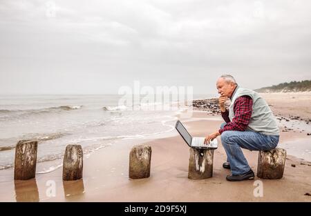 Un homme âgé attentionné avec un ordinateur portable est assis sur un fond marin. Travail pendant la quarantaine, pandémie Banque D'Images