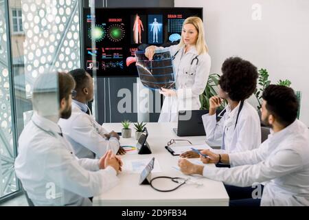 Jolie femme blonde d'âge moyen médecin a un discours à l'hôpital séminaire, se tient devant l'écran numérique avec des données, tient CT et la consulte Banque D'Images