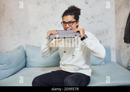 Jeune homme en colère avec des cheveux noirs bouclés va fou et en colère contre l'ordinateur portable Banque D'Images