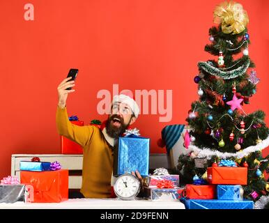 Le père Noël tient le téléphone cellulaire et la boîte à cadeaux. Un homme avec une barbe et un visage heureux envoie des salutations le nouvel an. Gadgets et concept de célébration. Le Père Noël prend le selfie près du sapin sur fond rouge Banque D'Images