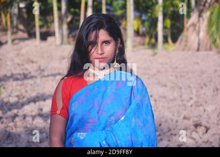 Gros plan d'une adolescente bengali indienne portant des boucles d'oreilles sari bleues collier blouse rouge avec de longs cheveux foncés debout dans un champ labouré. Banque D'Images