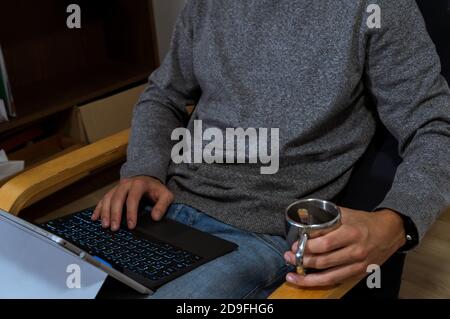 Jeune homme tenant un ordinateur portable sur les genoux, travaillant dessus, tapant dans un pull-over gris avec un café Banque D'Images