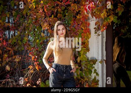 Une fille aux cheveux longs dans un body et un Jean posant dans un parc d'automne sur le fond d'un mur surcultivé avec des raisins sauvages. Filles dans le parc Banque D'Images