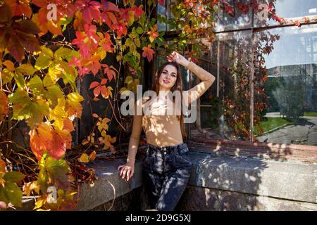 Une fille aux cheveux longs dans un body et un Jean posant dans un parc d'automne sur le fond d'un mur surcultivé avec des raisins sauvages. Filles dans le parc Banque D'Images