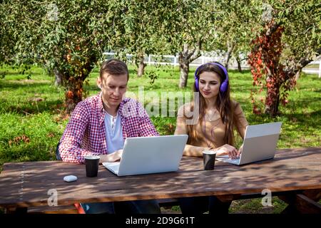 Un gars et une fille s'assoient à une table en bois dans le parc et travaillent sur des ordinateurs portables avec des écouteurs. Freelance travaille à l'extérieur. Travailler à l'extérieur du bureau en raison de Banque D'Images