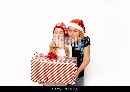 Maman et fille choquée, surprise et heureuse en chapeaux de Père Noël, tenant un grand cadeau de Noël dans leurs mains sur un fond blanc. Photo avec copie Banque D'Images