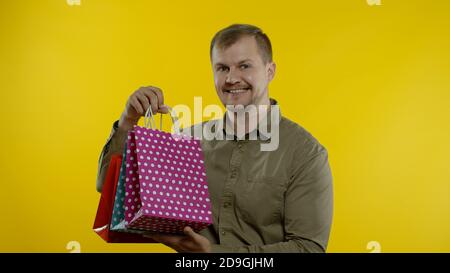 Joyeux homme montrant l'inscription du Vendredi fou de sacs de shopping, souriant regardant satisfait avec les bas prix, shopping le grand jour de vente. Studio tourné sur fond jaune Banque D'Images