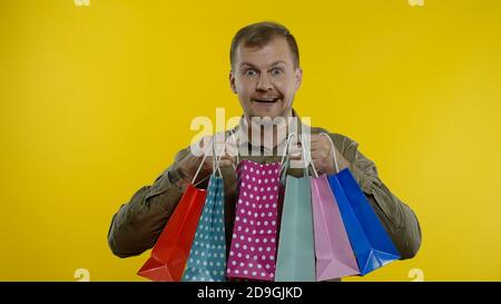 Joyeux homme montrant l'inscription du Vendredi fou de sacs de shopping, souriant regardant satisfait avec les bas prix, shopping le grand jour de vente. Studio tourné sur fond jaune Banque D'Images