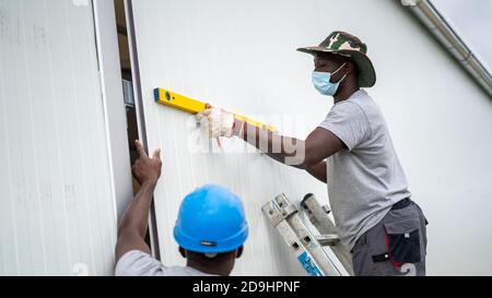 Constructeur afro-américain portant un masque facial et mesurant la longueur du mur Banque D'Images