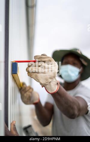 Constructeur afro-américain portant un masque facial et mesurant la longueur du mur Banque D'Images