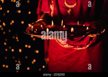 Femme indienne ou mariée portant une robe rouge traditionnelle et des bijoux, tenant puja thaali plein de lampes diya ou argile illuminées, célébrant Diwali la nuit. Banque D'Images