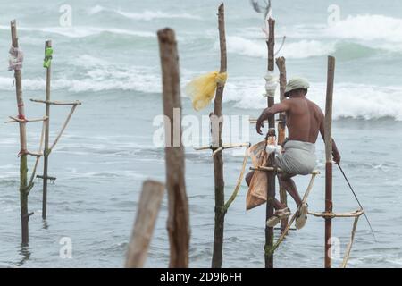 Vieux pêcheur de pilotis avec sa tige en bois faisant face à l'ange de caméra, la pêche dans une méthode traditionnelle unique dans la culture sri lankaise, lumière vive de evenin Banque D'Images