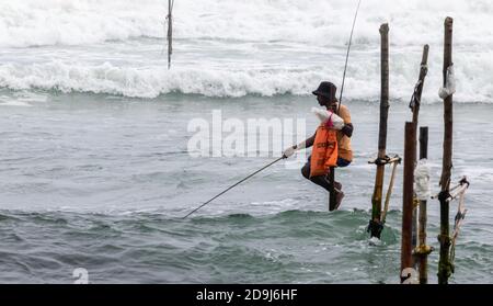 Weligama, province du Sud / Sri Lanka - 07 26 2020: Pêcheurs de pilotis avec sa tige en bois face à l'appareil photo avec sa poche orange, pêche dans Banque D'Images