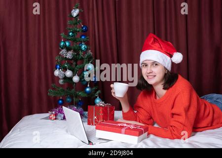Une jeune femme dans un chapeau de père Noël et un chandail rouge tient une tasse de café ou de thé, repose avec son ordinateur portable, allongé sur le lit, entouré de cadeaux de Noël. Banque D'Images