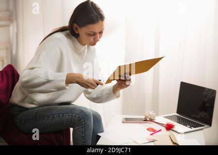 Confortable. Une femme ouvre, reçoit une carte de vœux pour le nouvel an et Noël 2021 de la part d'amis ou de la famille assis à la maison. Ouverture de l'enveloppe à l'aide de ciseaux. Vacances, fête. Il a l'air gai. Banque D'Images