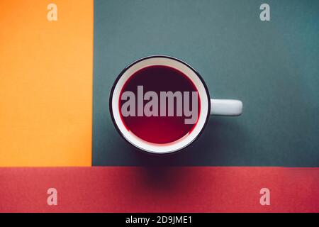 Une tasse de thé rouge Hibiscus sur fond carré blanc coloré dans des couleurs chaudes, orange terre cuite, vert foncé et jaune. Banque D'Images